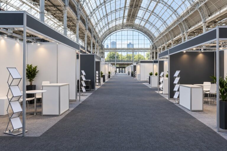 An empty exhibition hall aisle lined with blank modular stands under a glazed roof, before the doors open
